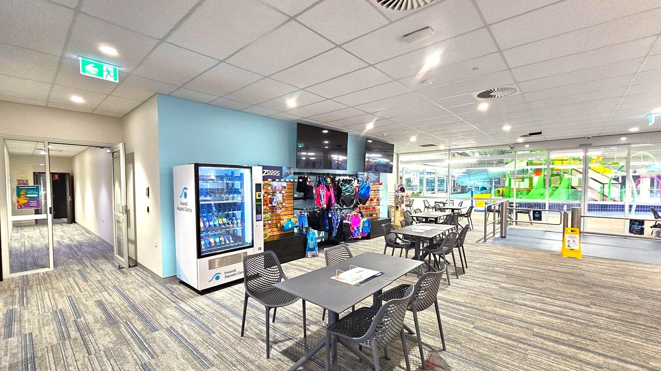 A room with a vending machine on the left, next to a display of swimwear and accessories. In the centre, there are tables and chairs with a magazine on one table. The floor is carpeted with a striped pattern. On the right, large glass windows reveal a view of an indoor pool area with colourful slides. The ceiling is fitted with recessed lighting and air vents. An exit sign is visible above a door on the left. A room with a vending machine on the left, next to a display of swimwear and accessories. In the centre, there are tables and chairs with a magazine on one table. The floor is carpeted with a striped pattern. On the right, large glass windows reveal a view of an indoor pool area with colourful slides. The ceiling is fitted with recessed lighting and air vents. An exit sign is visible above a door on the left.