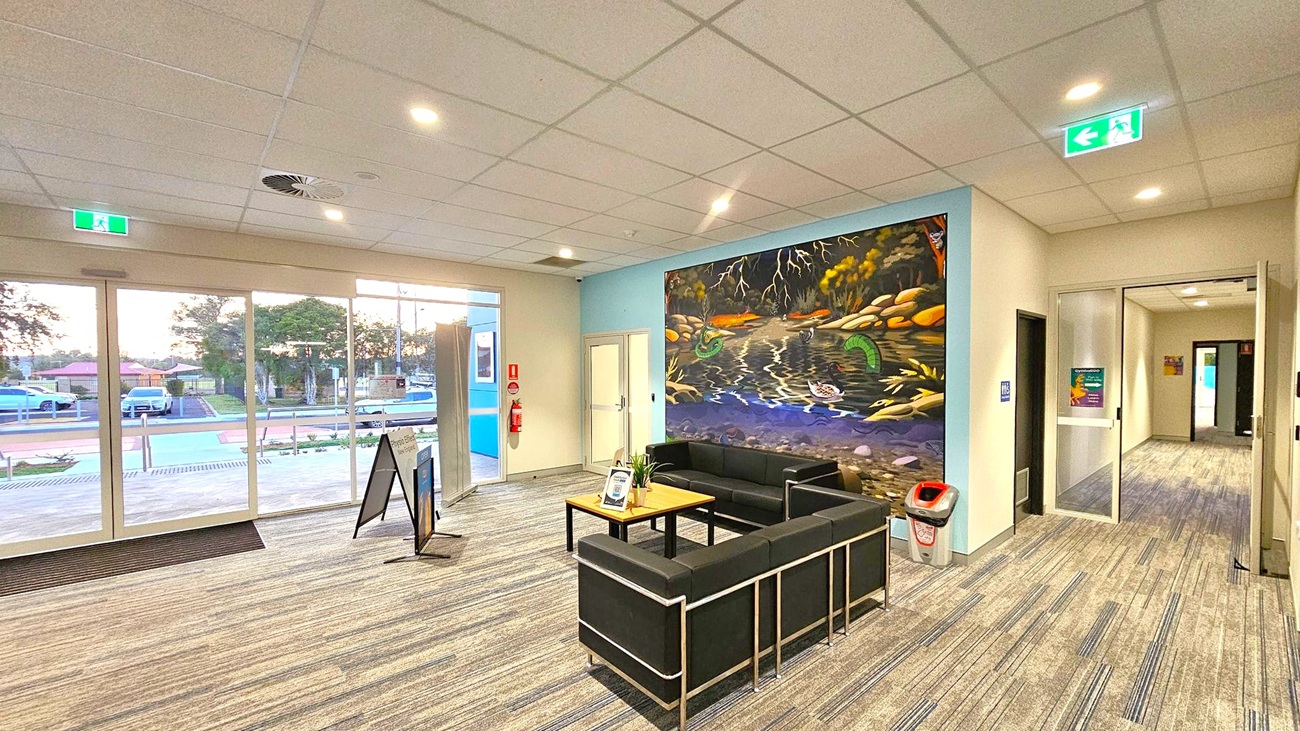 A spacious indoor area with a patterned carpet floor and a grid-patterned ceiling with recessed lights. On the left, large glass doors and windows reveal a view of parked cars and trees outside. In the centre, a seating area with black sofas and a wooden table. A vibrant mural of a river scene with rocks and foliage adorns the wall behind the sofas. On the right, a hallway leads further into the building, and a green exit sign is visible above. A fire extinguisher and a bin are near the mural. A spacious indoor area with a patterned carpet floor and a grid-patterned ceiling with recessed lights. On the left, large glass doors and windows reveal a view of parked cars and trees outside. In the centre, a seating area with black sofas and a wooden table. A vibrant mural of a river scene with rocks and foliage adorns the wall behind the sofas. On the right, a hallway leads further into the building, and a green exit sign is visible above. A fire extinguisher and a bin are near the mural.