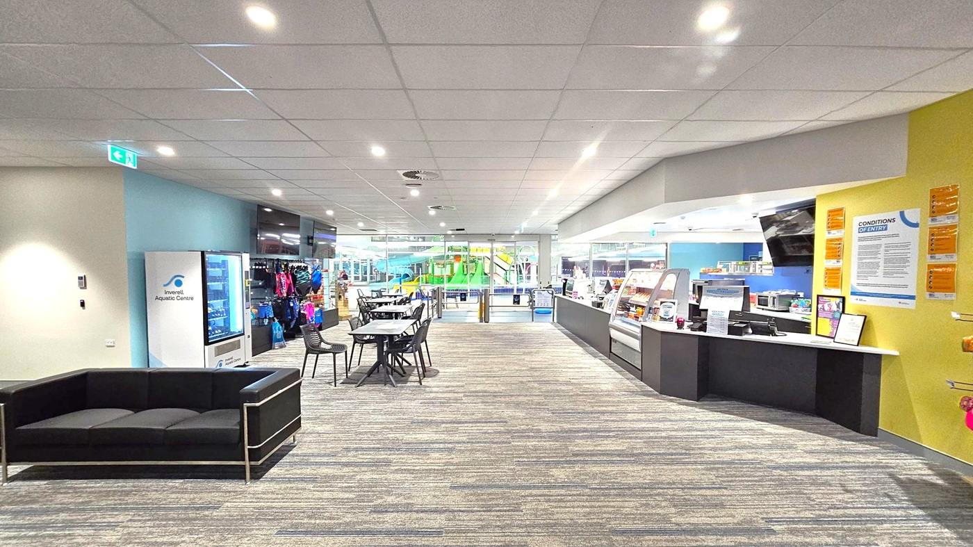 A spacious indoor area with a patterned carpet floor and a tiled ceiling with recessed lighting. On the left, there's a black sofa and a vending machine labelled "Inverell Aquatic Centre." In the centre, several tables and chairs are arranged. On the right, a counter with a display case and various notices on a yellow wall. In the background, large glass windows reveal a view of a pool area with green water slides. A spacious indoor area with a patterned carpet floor and a tiled ceiling with recessed lighting. On the left, there's a black sofa and a vending machine labelled "Inverell Aquatic Centre." In the centre, several tables and chairs are arranged. On the right, a counter with a display case and various notices on a yellow wall. In the background, large glass windows reveal a view of a pool area with green water slides.