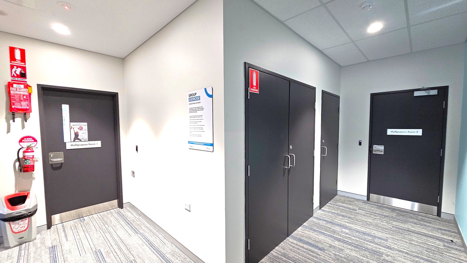 A hallway with grey carpet and a white ceiling featuring recessed lights. On the left, a black door labelled "Multipurpose Room 1" is accompanied by a fire extinguisher, fire blanket, and a bin. A sign about group exercise is mounted on the wall. On the right, two black doors are visible, one labelled "Multipurpose Room 2" and another unlabelled. Both sides have emergency exit signs above the doors. A hallway with grey carpet and a white ceiling featuring recessed lights. On the left, a black door labelled "Multipurpose Room 1" is accompanied by a fire extinguisher, fire blanket, and a bin. A sign about group exercise is mounted on the wall. On the right, two black doors are visible, one labelled "Multipurpose Room 2" and another unlabelled. Both sides have emergency exit signs above the doors.