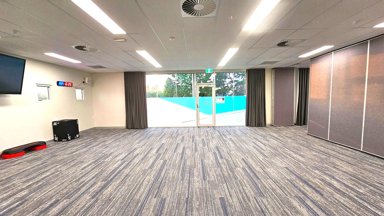 A spacious room with a grey carpet featuring subtle coloured stripes. The ceiling has recessed lighting and air vents. On the left, a wall-mounted digital clock displays "6:25" in red and blue. Below it, a black exercise box and red step platforms are visible. The back wall has large glass doors with a blue and white design, flanked by dark curtains. On the right, there are large, foldable partition panels. A spacious room with a grey carpet featuring subtle coloured stripes. The ceiling has recessed lighting and air vents. On the left, a wall-mounted digital clock displays "6:25" in red and blue. Below it, a black exercise box and red step platforms are visible. The back wall has large glass doors with a blue and white design, flanked by dark curtains. On the right, there are large, foldable partition panels.