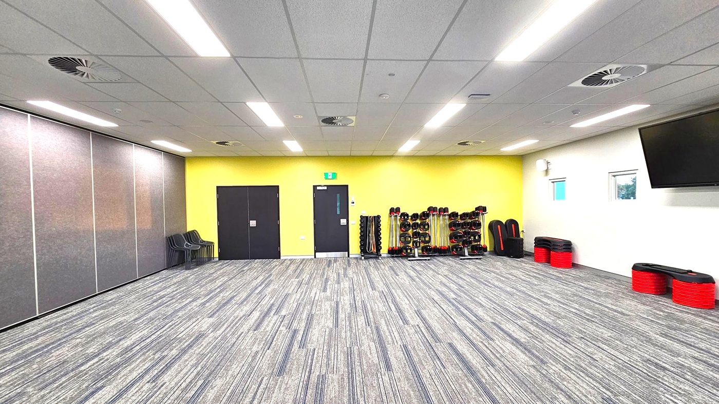 A spacious room with a grey carpeted floor and a ceiling with fluorescent lights and air vents. On the left, there are grey partition walls and a stack of black chairs. The back wall is yellow with two black doors. In the centre, there are racks of weights and exercise equipment. On the right, a white wall features two small windows and a large TV screen. Red and black exercise steps are stacked against the wall. A spacious room with a grey carpeted floor and a ceiling with fluorescent lights and air vents. On the left, there are grey partition walls and a stack of black chairs. The back wall is yellow with two black doors. In the centre, there are racks of weights and exercise equipment. On the right, a white wall features two small windows and a large TV screen. Red and black exercise steps are stacked against the wall.