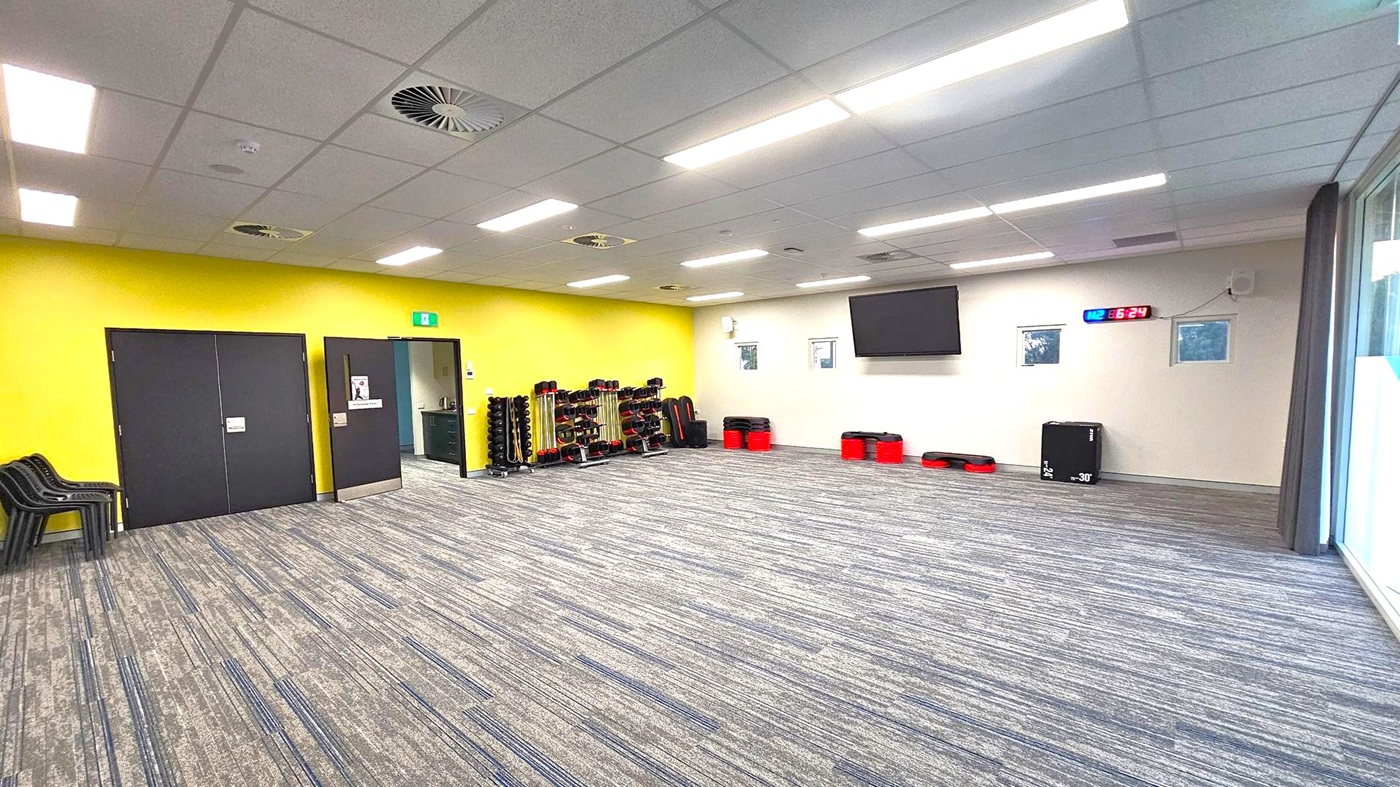 A spacious room with a grey carpeted floor and a ceiling with fluorescent lights and air vents. On the left, there are black double doors and a stack of black chairs. In the middle, a yellow wall features racks of weights and exercise equipment. On the right, a white wall has a large TV screen, a digital clock displaying "16:24", and small windows. Red and black exercise steps and a black box are placed against the wall. Large windows with curtains are on the far right. A spacious room with a grey carpeted floor and a ceiling with fluorescent lights and air vents. On the left, there are black double doors and a stack of black chairs. In the middle, a yellow wall features racks of weights and exercise equipment. On the right, a white wall has a large TV screen, a digital clock displaying "16:24", and small windows. Red and black exercise steps and a black box are placed against the wall. Large windows with curtains are on the far right.