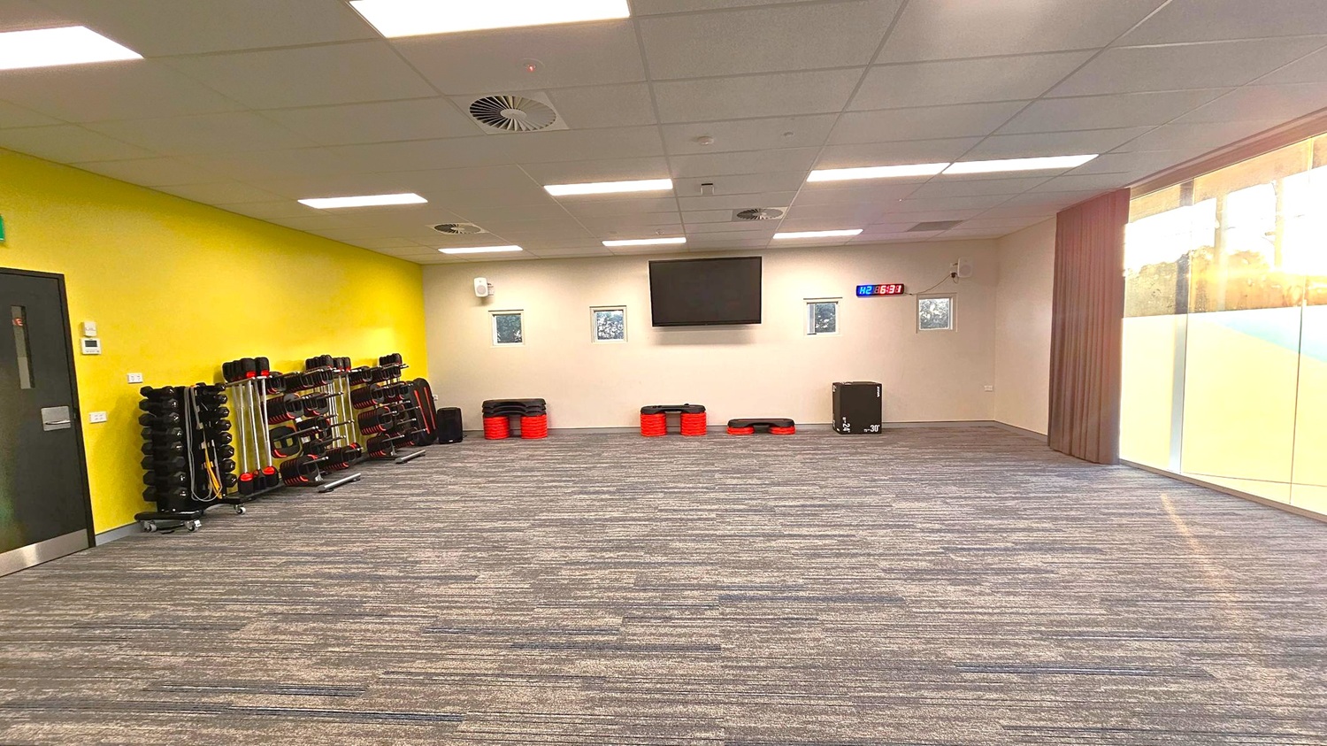 A spacious room with a grey and purple carpeted floor. On the left, a yellow wall features a black door and racks of fitness equipment, including weights and steps. The back wall is white with a large TV screen and a digital clock displaying "6:31". Small windows are positioned on either side of the TV. On the right, large glass windows allow natural light to enter, with a view of trees outside. The ceiling is fitted with white panels and fluorescent lights. A spacious room with a grey and purple carpeted floor. On the left, a yellow wall features a black door and racks of fitness equipment, including weights and steps. The back wall is white with a large TV screen and a digital clock displaying "6:31". Small windows are positioned on either side of the TV. On the right, large glass windows allow natural light to enter, with a view of trees outside. The ceiling is fitted with white panels and fluorescent lights.