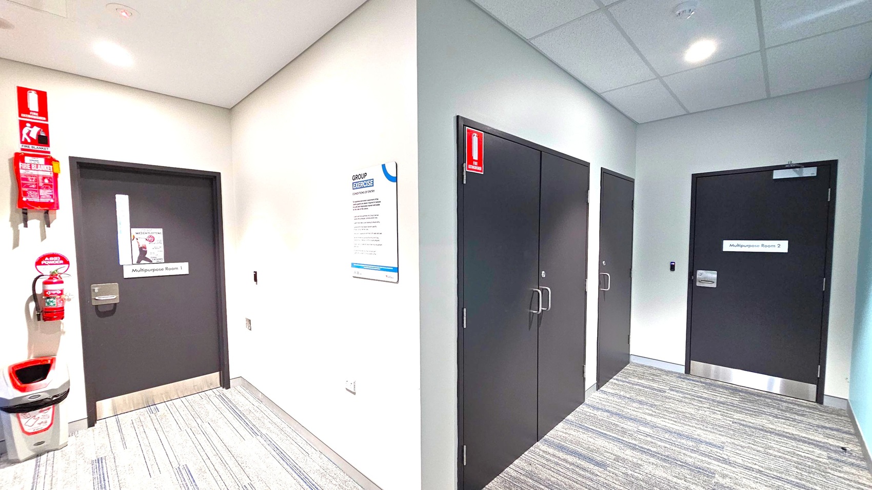 A hallway with grey carpet and white walls. On the left, a door labelled "Multipurpose Room 1" is next to a fire extinguisher and a fire blanket mounted on the wall. A bin is positioned below them. In the centre, a sign titled "Group Exercise" is affixed to the wall. On the right, there are two black doors, one labelled "Multipurpose Room 2". The ceiling is fitted with recessed lighting and a smoke detector. A hallway with grey carpet and white walls. On the left, a door labelled "Multipurpose Room 1" is next to a fire extinguisher and a fire blanket mounted on the wall. A bin is positioned below them. In the centre, a sign titled "Group Exercise" is affixed to the wall. On the right, there are two black doors, one labelled "Multipurpose Room 2". The ceiling is fitted with recessed lighting and a smoke detector.