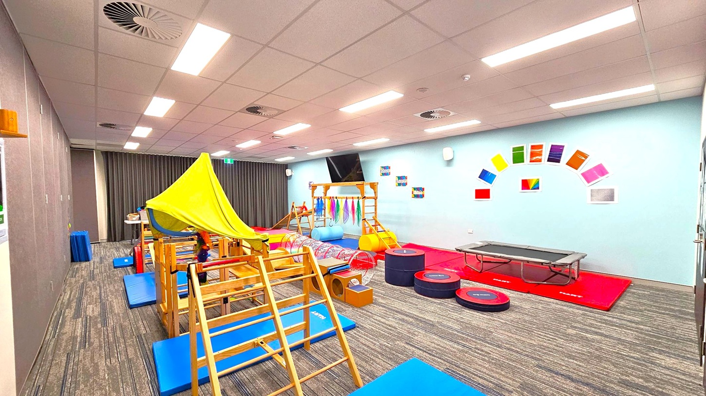 A children's playroom with a variety of equipment. On the left, there's a wooden climbing frame with a green fabric canopy. In the centre, colourful mats and a tunnel are visible. On the right, a trampoline is placed on red mats. The walls are light blue with colourful paper squares arranged in an arc. The floor is carpeted with a striped pattern, and the ceiling has recessed lighting and air vents. A children's playroom with a variety of equipment. On the left, there's a wooden climbing frame with a green fabric canopy. In the centre, colourful mats and a tunnel are visible. On the right, a trampoline is placed on red mats. The walls are light blue with colourful paper squares arranged in an arc. The floor is carpeted with a striped pattern, and the ceiling has recessed lighting and air vents.
