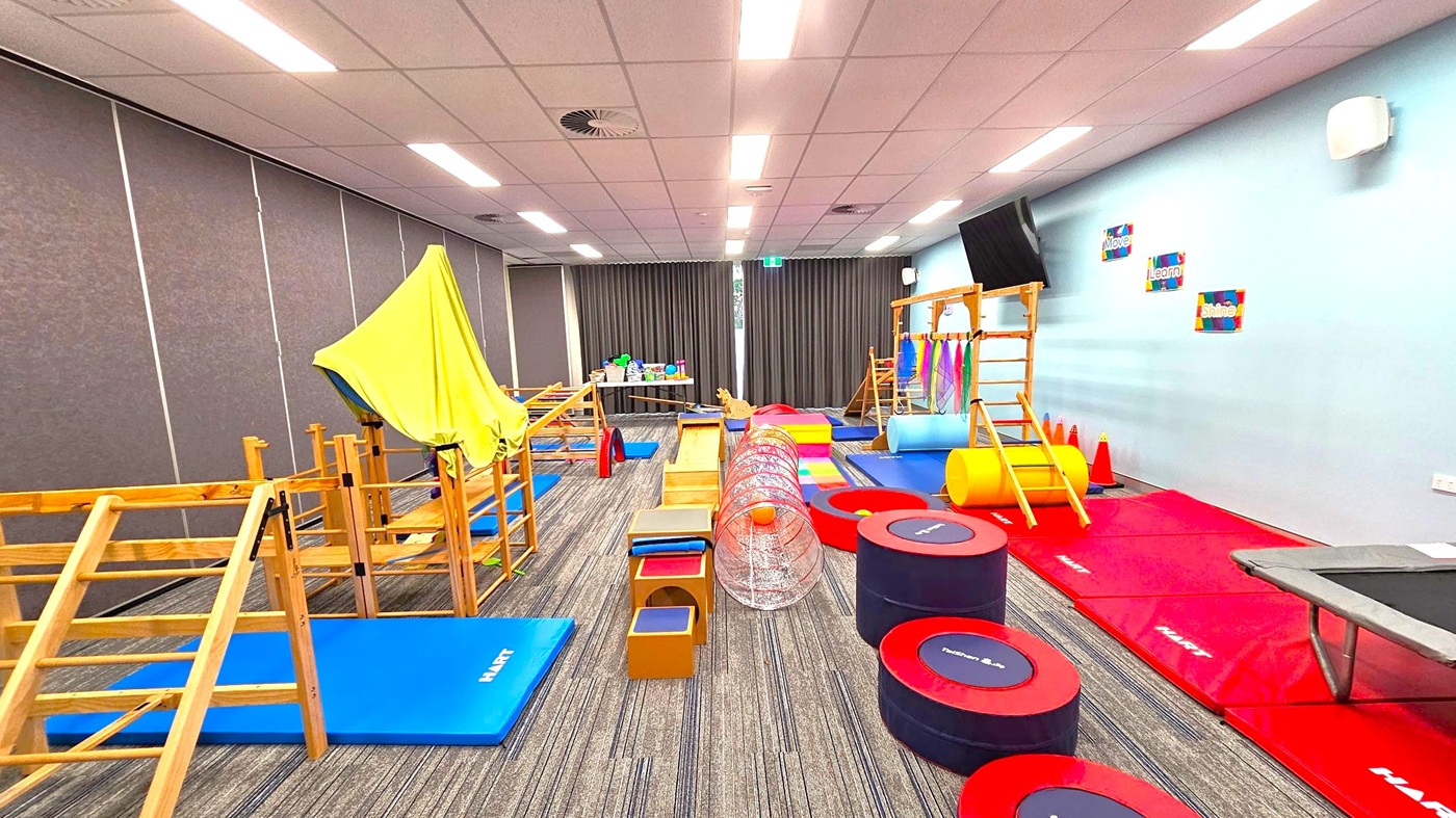 A room with a variety of gym equipment for children. On the left, there are wooden climbing structures and blue mats. In the centre, a tunnel and various soft play items are arranged on a carpeted floor. On the right, red mats, a trampoline, and a wooden ladder are visible. The walls are light blue with motivational posters, and the ceiling has fluorescent lights. A table with toys is at the back near dark curtains. A room with a variety of gym equipment for children. On the left, there are wooden climbing structures and blue mats. In the centre, a tunnel and various soft play items are arranged on a carpeted floor. On the right, red mats, a trampoline, and a wooden ladder are visible. The walls are light blue with motivational posters, and the ceiling has fluorescent lights. A table with toys is at the back near dark curtains.