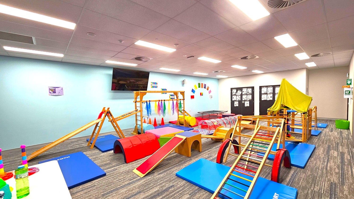 A children's playroom with various wooden climbing structures and colourful mats on the floor. On the left, a table holds stacked toys and bottles. In the centre, a wooden frame with hanging scarves and a slide is surrounded by red and blue mats. On the right, a structure is covered with a green cloth. The walls are light blue with colourful decorations, and a large TV is mounted on the left wall. The ceiling has recessed lighting, and the floor is carpeted with a striped pattern. A children's playroom with various wooden climbing structures and colourful mats on the floor. On the left, a table holds stacked toys and bottles. In the centre, a wooden frame with hanging scarves and a slide is surrounded by red and blue mats. On the right, a structure is covered with a green cloth. The walls are light blue with colourful decorations, and a large TV is mounted on the left wall. The ceiling has recessed lighting, and the floor is carpeted with a striped pattern.