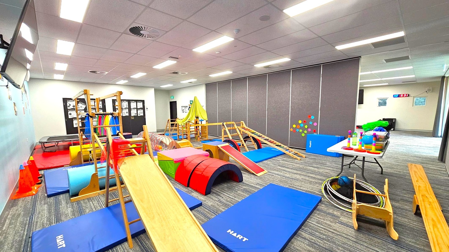 A children's playroom with various gym equipment and toys. On the left, there are wooden climbing structures, colourful mats, and cones. In the centre, a slide and a tunnel are surrounded by soft play shapes. On the right, a table holds stacking toys and other items, with hula hoops and a small wooden chair nearby. The floor is carpeted with a striped pattern, and the ceiling has recessed lighting and ventilation. Walls are decorated with colourful dots and posters. A children's playroom with various gym equipment and toys. On the left, there are wooden climbing structures, colourful mats, and cones. In the centre, a slide and a tunnel are surrounded by soft play shapes. On the right, a table holds stacking toys and other items, with hula hoops and a small wooden chair nearby. The floor is carpeted with a striped pattern, and the ceiling has recessed lighting and ventilation. Walls are decorated with colourful dots and posters.