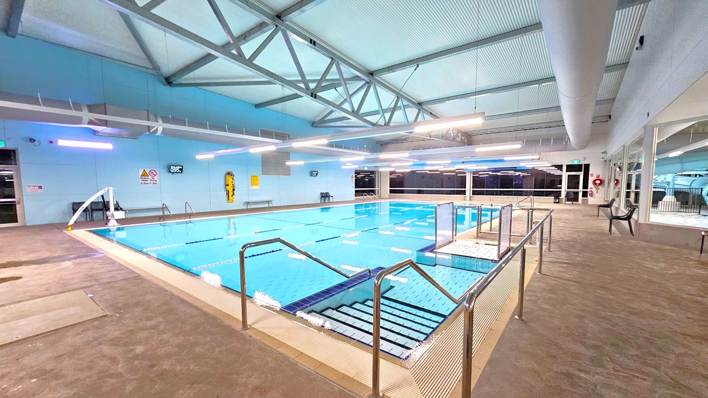 An indoor swimming pool with clear blue water, surrounded by a beige tiled deck. Stainless steel handrails lead into the pool. On the left, a wall with safety signs and a digital clock. The ceiling features exposed metal beams and fluorescent lights. On the right, large windows and glass doors, with a few plastic chairs along the wall. The floor is a textured surface, and the ceiling is lined with ductwork. An indoor swimming pool with clear blue water, surrounded by a beige tiled deck. Stainless steel handrails lead into the pool. On the left, a wall with safety signs and a digital clock. The ceiling features exposed metal beams and fluorescent lights. On the right, large windows and glass doors, with a few plastic chairs along the wall. The floor is a textured surface, and the ceiling is lined with ductwork.