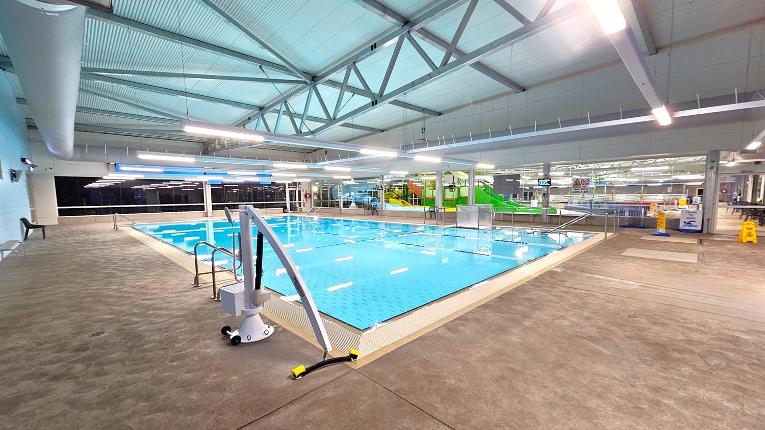 Indoor swimming pool area with a clear blue pool in the centre. The pool is surrounded by a beige tiled floor. On the left, there is a chair against the wall. In the background, large windows and a green water slide are visible. The ceiling is high with exposed beams and fluorescent lights. On the right, there are caution signs and a seating area with tables and chairs. Indoor swimming pool area with a clear blue pool in the centre. The pool is surrounded by a beige tiled floor. On the left, there is a chair against the wall. In the background, large windows and a green water slide are visible. The ceiling is high with exposed beams and fluorescent lights. On the right, there are caution signs and a seating area with tables and chairs.