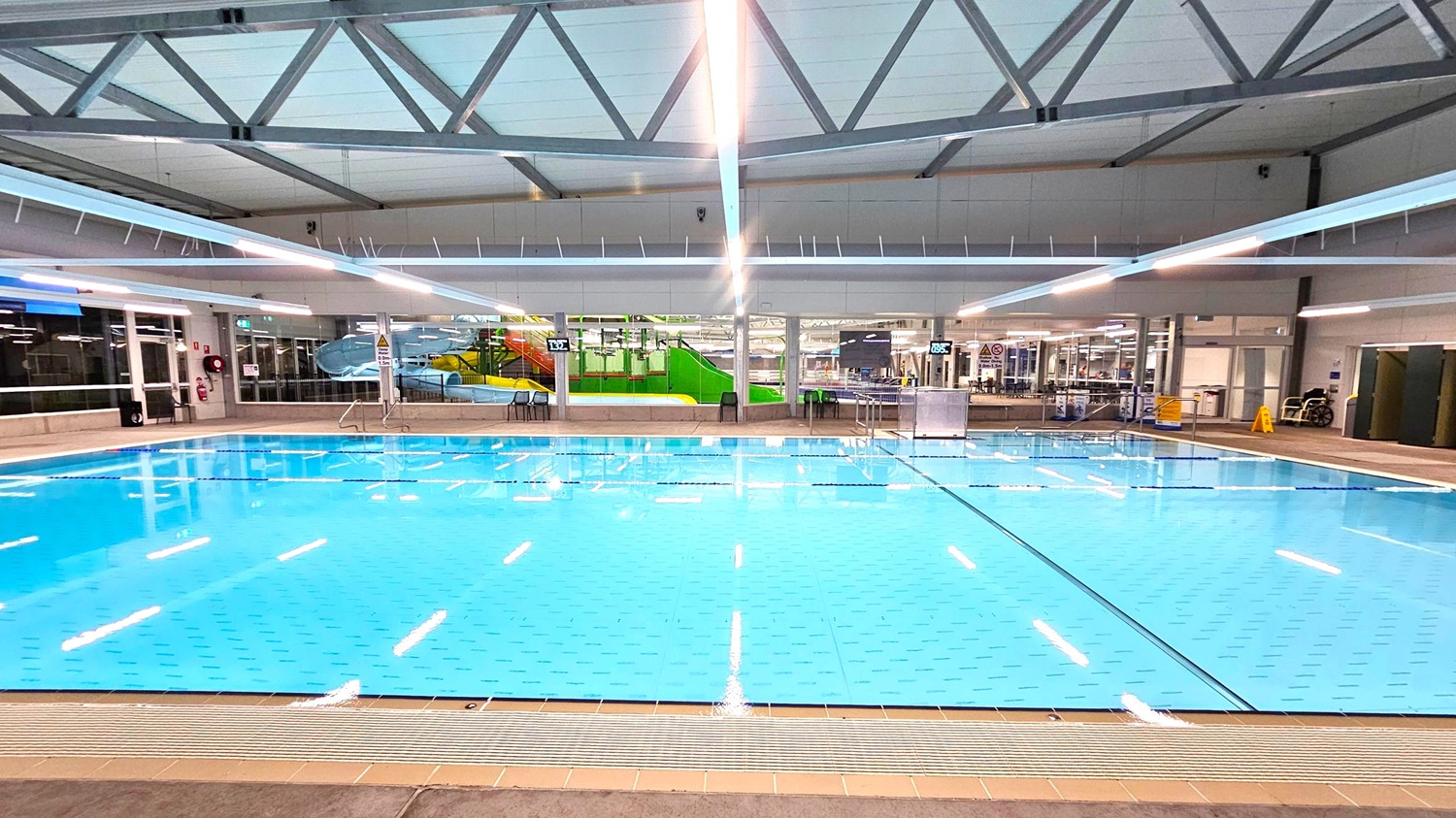 An indoor swimming pool with clear blue water, surrounded by a tiled deck. In the background, there are large windows and a colourful water slide structure with green and yellow elements. The ceiling features exposed metal beams and bright overhead lights. On the right, there are some chairs and a caution sign. The pool area is spacious and well-lit. An indoor swimming pool with clear blue water, surrounded by a tiled deck. In the background, there are large windows and a colourful water slide structure with green and yellow elements. The ceiling features exposed metal beams and bright overhead lights. On the right, there are some chairs and a caution sign. The pool area is spacious and well-lit.