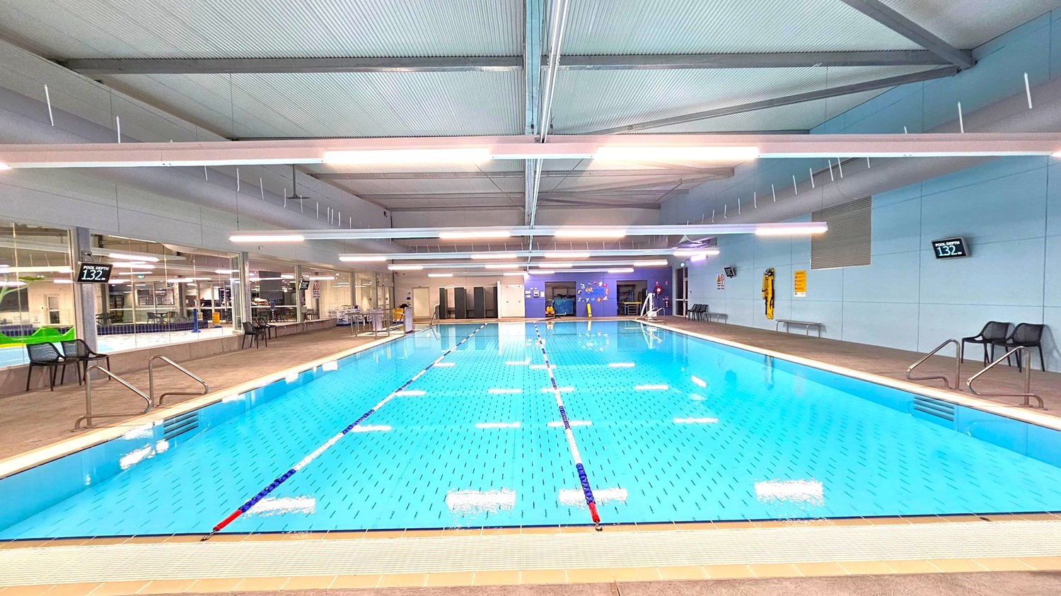 An indoor swimming pool with clear blue water, marked with lane dividers. On the left, there are chairs and a digital display showing the pool depth as 1.32 metres. The right side also has chairs and a similar depth display. The background features large windows, tables, and a wall with safety equipment. The ceiling is high with exposed beams and fluorescent lighting. The floor around the pool is tiled. An indoor swimming pool with clear blue water, marked with lane dividers. On the left, there are chairs and a digital display showing the pool depth as 1.32 metres. The right side also has chairs and a similar depth display. The background features large windows, tables, and a wall with safety equipment. The ceiling is high with exposed beams and fluorescent lighting. The floor around the pool is tiled.