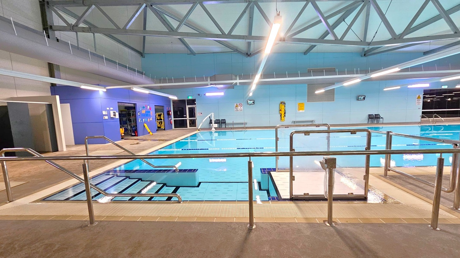 Indoor swimming pool with metal railings in the foreground. Steps lead into the pool on the left, and a hoist is positioned on the right. The pool area is surrounded by a tiled floor. The back wall is blue with various signs and a digital clock. Overhead, there are exposed metal beams and fluorescent lights. A door and some equipment are visible on the left wall. Indoor swimming pool with metal railings in the foreground. Steps lead into the pool on the left, and a hoist is positioned on the right. The pool area is surrounded by a tiled floor. The back wall is blue with various signs and a digital clock. Overhead, there are exposed metal beams and fluorescent lights. A door and some equipment are visible on the left wall.
