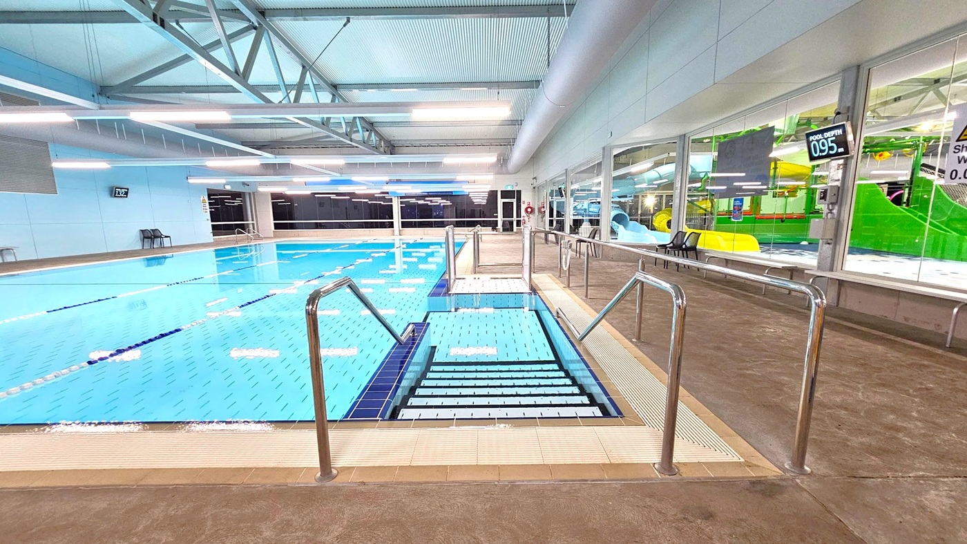 Indoor swimming pool with clear blue water and stainless steel handrails leading into the pool. On the left, the pool extends with lane dividers and a depth sign of 1.32 metres. On the right, a glass wall reveals a play area with colourful slides and structures. The floor is tiled, and the ceiling features exposed beams and bright lighting. A depth sign of 0.95 metres is visible on the right wall. Indoor swimming pool with clear blue water and stainless steel handrails leading into the pool. On the left, the pool extends with lane dividers and a depth sign of 1.32 metres. On the right, a glass wall reveals a play area with colourful slides and structures. The floor is tiled, and the ceiling features exposed beams and bright lighting. A depth sign of 0.95 metres is visible on the right wall.