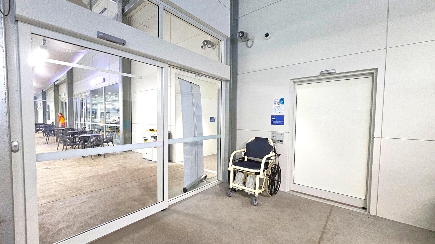 On the left, there is a glass sliding door leading to an indoor area with tables and chairs. A person in a red and yellow outfit is visible in the background. On the right, a white door with a sign indicating an accessible adult changing facility is present. A wheelchair is positioned next to the door. The floor is a mix of beige and green, and the ceiling is white with fluorescent lighting. Security cameras are mounted on the wall above the door. On the left, there is a glass sliding door leading to an indoor area with tables and chairs. A person in a red and yellow outfit is visible in the background. On the right, a white door with a sign indicating an accessible adult changing facility is present. A wheelchair is positioned next to the door. The floor is a mix of beige and green, and the ceiling is white with fluorescent lighting. Security cameras are mounted on the wall above the door.