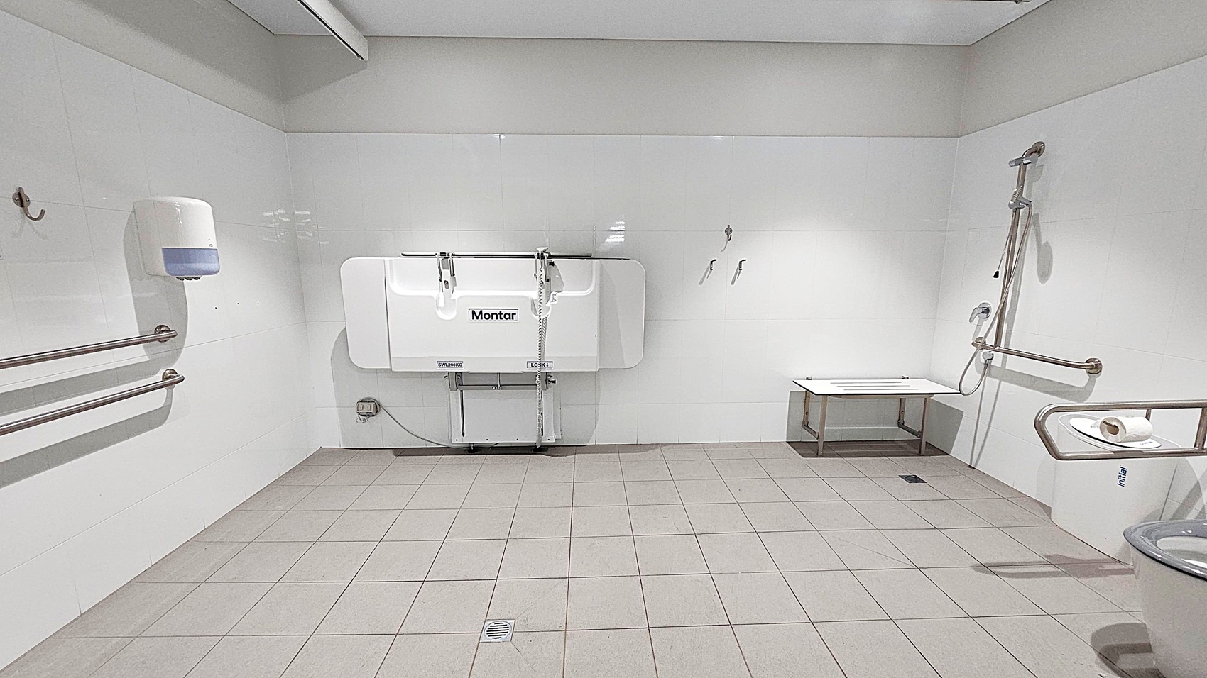 A spacious, accessible bathroom with grey tiled flooring and white walls. On the left, there are horizontal metal grab bars and a wall-mounted soap dispenser. In the centre, a foldable changing table is attached to the wall. On the right, a shower area with a handheld showerhead and a foldable seat is visible. A toilet with a grab bar and a toilet roll holder is in the foreground. The ceiling is plain with a light fixture. A spacious, accessible bathroom with grey tiled flooring and white walls. On the left, there are horizontal metal grab bars and a wall-mounted soap dispenser. In the centre, a foldable changing table is attached to the wall. On the right, a shower area with a handheld showerhead and a foldable seat is visible. A toilet with a grab bar and a toilet roll holder is in the foreground. The ceiling is plain with a light fixture.