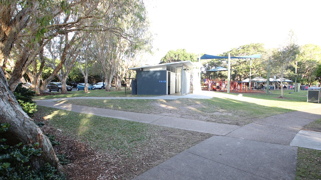 In the foreground, a paved pathway intersects with a grassy area. On the left, several trees provide shade, and a row of parked cars is visible behind them. To the right, a modern grey building features a small roof overhang and is positioned adjacent to a playground with multiple play structures, including slides and climbing equipment. The ground consists of smooth concrete for the path and soft grass in the surrounding area, creating a functional space for both play and relaxation. In the foreground, a paved pathway intersects with a grassy area. On the left, several trees provide shade, and a row of parked cars is visible behind them. To the right, a modern grey building features a small roof overhang and is positioned adjacent to a playground with multiple play structures, including slides and climbing equipment. The ground consists of smooth concrete for the path and soft grass in the surrounding area, creating a functional space for both play and relaxation.