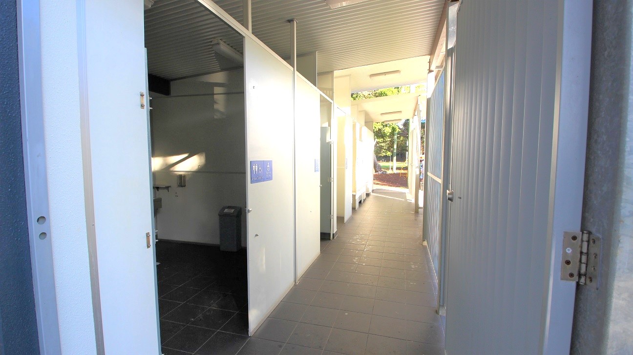 In the foreground, a narrow corridor is lined with individual restroom cubicles on either side. To the left, the doors are installed against a light-coloured wall, while on the right, there are grey panel doors leading to the facilities. The floor is covered with dark tiles, and the ceiling has fluorescent lighting, providing ample illumination. A waste bin is visible in one of the cubicles, and sunlight streams in through the entrance. In the foreground, a narrow corridor is lined with individual restroom cubicles on either side. To the left, the doors are installed against a light-coloured wall, while on the right, there are grey panel doors leading to the facilities. The floor is covered with dark tiles, and the ceiling has fluorescent lighting, providing ample illumination. A waste bin is visible in one of the cubicles, and sunlight streams in through the entrance.
