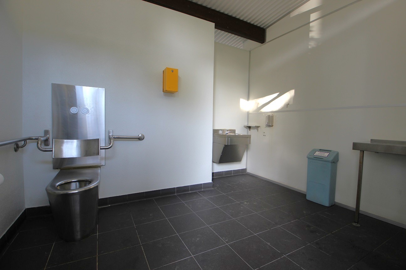 In the foreground, a restroom area features a stainless steel toilet on the left, equipped with grab rails for accessibility. To the right, a sink is mounted against the wall, with a small soap dispenser above it. A light blue trash bin is positioned nearby. On the wall, an orange box is mounted, likely containing supplies. The floor is covered with dark tiles, while the ceiling is lined with metal panels, enhancing the clean and functional design of the space. In the foreground, a restroom area features a stainless steel toilet on the left, equipped with grab rails for accessibility. To the right, a sink is mounted against the wall, with a small soap dispenser above it. A light blue trash bin is positioned nearby. On the wall, an orange box is mounted, likely containing supplies. The floor is covered with dark tiles, while the ceiling is lined with metal panels, enhancing the clean and functional design of the space.