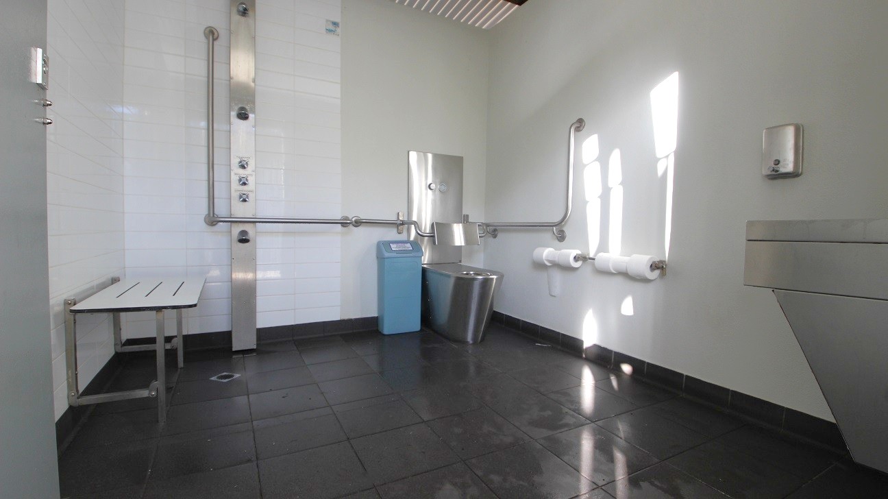 In the foreground, a clean restroom area features a stainless steel sink on the right, with a mounted soap dispenser above it. Nearby, a light blue waste bin is positioned against the wall, alongside two rolls of toilet paper affixed to the wall. A folding white bench is located to the left, supported by pipes. Vertical and horizontal metal grab rails are installed along the wall for accessibility. The floor is tiled with dark tiles, while the ceiling is lined with metal panels. In the foreground, a clean restroom area features a stainless steel sink on the right, with a mounted soap dispenser above it. Nearby, a light blue waste bin is positioned against the wall, alongside two rolls of toilet paper affixed to the wall. A folding white bench is located to the left, supported by pipes. Vertical and horizontal metal grab rails are installed along the wall for accessibility. The floor is tiled with dark tiles, while the ceiling is lined with metal panels.