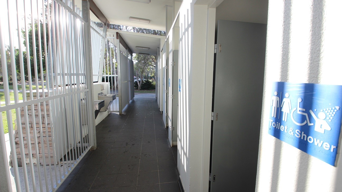 In the foreground, a corridor leads to restroom facilities, with metal railings along the left side. A sink area is built into the wall, providing washing facilities. On the right, several restroom cubicle doors are visible, with one showing a sign indicating "Toilet & Shower" in blue. The floor is tiled with dark tiles, while the ceiling features overhead lighting that adds to the brightness of the space. In the foreground, a corridor leads to restroom facilities, with metal railings along the left side. A sink area is built into the wall, providing washing facilities. On the right, several restroom cubicle doors are visible, with one showing a sign indicating "Toilet & Shower" in blue. The floor is tiled with dark tiles, while the ceiling features overhead lighting that adds to the brightness of the space.