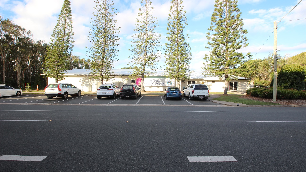 A row of parked cars is in front of a single-storey building with a light-coloured exterior. Tall, narrow trees are evenly spaced along the front of the building. The foreground features a multi-lane road with visible lane markings. On the left, there are more trees, while on the right, there is a grassy area with bushes and a utility pole. A row of parked cars is in front of a single-storey building with a light-coloured exterior. Tall, narrow trees are evenly spaced along the front of the building. The foreground features a multi-lane road with visible lane markings. On the left, there are more trees, while on the right, there is a grassy area with bushes and a utility pole.