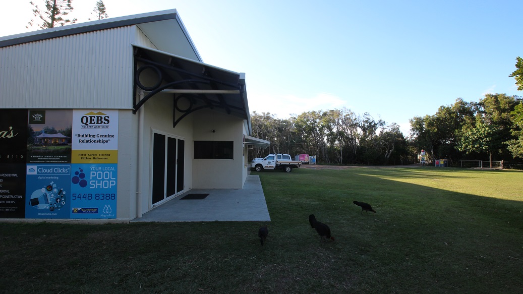 A building with a corrugated metal exterior is on the left, featuring several signs and advertisements. A small awning extends over the entrance. To the right, a grassy area stretches out, with a white utility vehicle parked in the distance. Three birds are visible on the grass. In the background, there are trees lining the edge of the open space. The ground is a mix of grass and dirt. A building with a corrugated metal exterior is on the left, featuring several signs and advertisements. A small awning extends over the entrance. To the right, a grassy area stretches out, with a white utility vehicle parked in the distance. Three birds are visible on the grass. In the background, there are trees lining the edge of the open space. The ground is a mix of grass and dirt.
