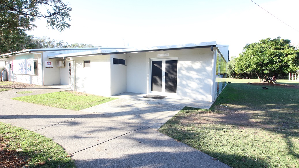 A small, single-storey white building with a flat roof is situated on a grassy area. On the right, there are double glass doors, and a narrow window is visible on the left side. A concrete path leads to the entrance, with grass on either side. In the background, there are trees and open space. Shadows from nearby trees are cast on the ground and building. A small, single-storey white building with a flat roof is situated on a grassy area. On the right, there are double glass doors, and a narrow window is visible on the left side. A concrete path leads to the entrance, with grass on either side. In the background, there are trees and open space. Shadows from nearby trees are cast on the ground and building.