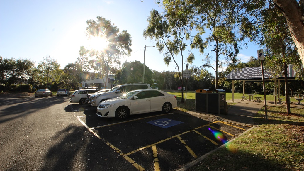 A car park with several vehicles, including a white car in a designated disabled parking space. The sun is low, casting long shadows and creating a lens flare. On the right, there are trees and a covered picnic area with tables. The ground is paved, and there is a grassy area adjacent to the parking lot. A car park with several vehicles, including a white car in a designated disabled parking space. The sun is low, casting long shadows and creating a lens flare. On the right, there are trees and a covered picnic area with tables. The ground is paved, and there is a grassy area adjacent to the parking lot.