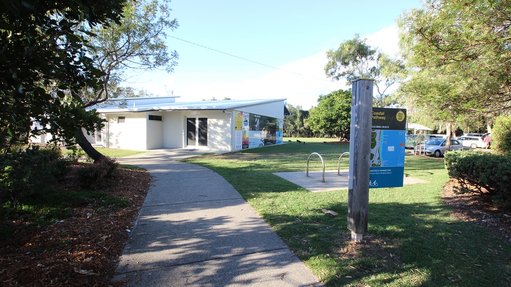 A pathway leads to a modern, single-storey building with large windows on the left. In the foreground, a signpost with a blue information board stands beside a bike rack. The area is surrounded by well-maintained grass and trees, with a few cars parked on the right. The scene is well-lit, suggesting daytime. A pathway leads to a modern, single-storey building with large windows on the left. In the foreground, a signpost with a blue information board stands beside a bike rack. The area is surrounded by well-maintained grass and trees, with a few cars parked on the right. The scene is well-lit, suggesting daytime.
