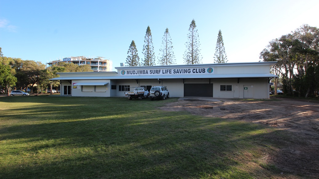 A single-storey building with a sign reading "Mudjimba Surf Life Saving Club" on the front. Two vehicles are parked in front of the building. The background features tall, narrow trees and a multi-storey building partially visible on the left. The foreground consists of a grassy area with patches of sunlight and shadow. Trees are visible on the right side. A single-storey building with a sign reading "Mudjimba Surf Life Saving Club" on the front. Two vehicles are parked in front of the building. The background features tall, narrow trees and a multi-storey building partially visible on the left. The foreground consists of a grassy area with patches of sunlight and shadow. Trees are visible on the right side.
