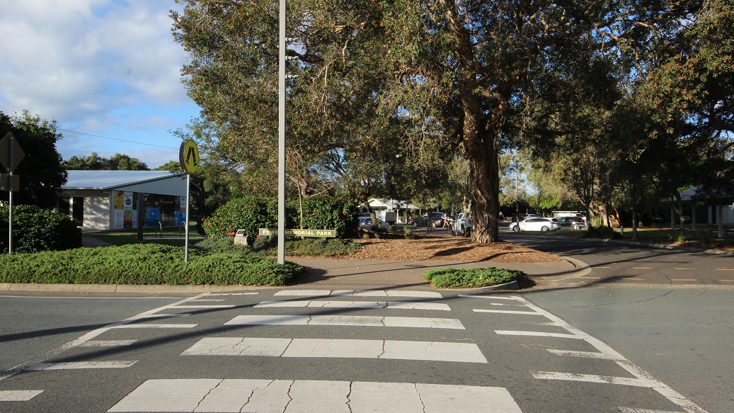 A zebra crossing leads to a path surrounded by greenery and a large tree in the centre. On the left, there's a building with a white roof partially visible behind bushes. A tall streetlight stands near the tree. In the background, several parked cars are visible among more trees. The road surface is smooth, and the area is well-lit, suggesting a sunny day. A zebra crossing leads to a path surrounded by greenery and a large tree in the centre. On the left, there's a building with a white roof partially visible behind bushes. A tall streetlight stands near the tree. In the background, several parked cars are visible among more trees. The road surface is smooth, and the area is well-lit, suggesting a sunny day.