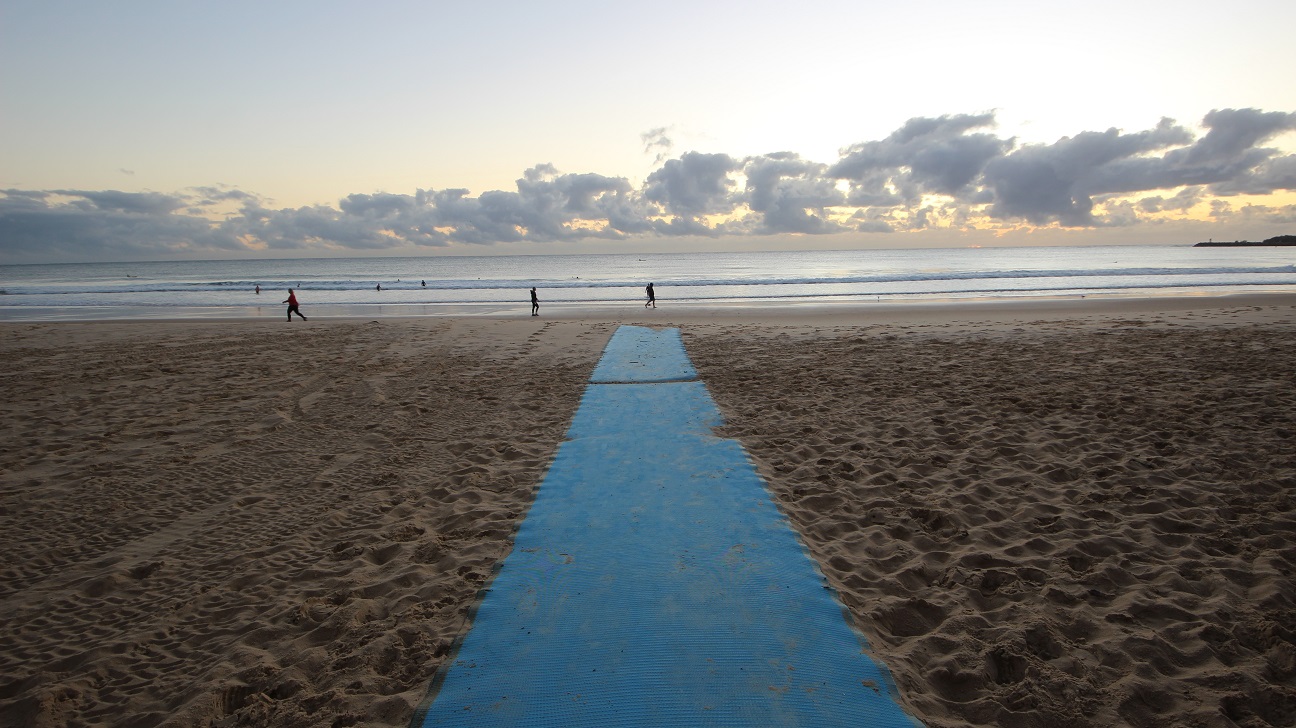 In the foreground, a blue mat runs across the sandy beach, leading towards the ocean. On the left, a person in a red shirt is walking along the beach, while in the distance, several people are wading in the water. To the right, another person is seen walking at the water's edge. The sand is finely textured, with some footprints visible leading towards the water. The scene conveys a calm beach environment, with the shoreline gently meeting the waves, creating a clear path for beachgoers. In the foreground, a blue mat runs across the sandy beach, leading towards the ocean. On the left, a person in a red shirt is walking along the beach, while in the distance, several people are wading in the water. To the right, another person is seen walking at the water's edge. The sand is finely textured, with some footprints visible leading towards the water. The scene conveys a calm beach environment, with the shoreline gently meeting the waves, creating a clear path for beachgoers.
