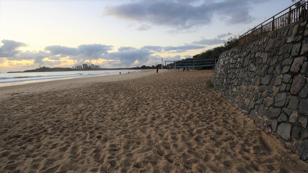 In the foreground, a sandy beach stretches out, with soft sand marked by footprints leading towards the ocean. On the left, a stone wall borders the beach, rising steadily and providing structural support. There are wooden railings on top of the wall, indicating a pathway or promenade above. In the distance, the coastline is visible, with several figures walking along the shore and a few people in the water. In the foreground, a sandy beach stretches out, with soft sand marked by footprints leading towards the ocean. On the left, a stone wall borders the beach, rising steadily and providing structural support. There are wooden railings on top of the wall, indicating a pathway or promenade above. In the distance, the coastline is visible, with several figures walking along the shore and a few people in the water.