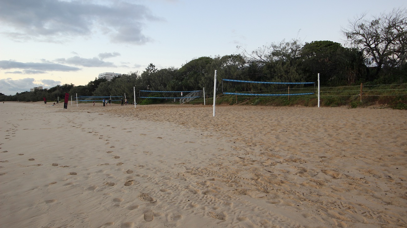 In the foreground, a sandy beach is visible, dotted with footprints that lead towards the water. On the left side, several red poles mark the boundaries of beach volleyball courts, while on the right, white posts with blue netting define additional courts. There are no people actively playing, but a few individuals can be seen in the distance engaged in activities near the courts. In the foreground, a sandy beach is visible, dotted with footprints that lead towards the water. On the left side, several red poles mark the boundaries of beach volleyball courts, while on the right, white posts with blue netting define additional courts. There are no people actively playing, but a few individuals can be seen in the distance engaged in activities near the courts.