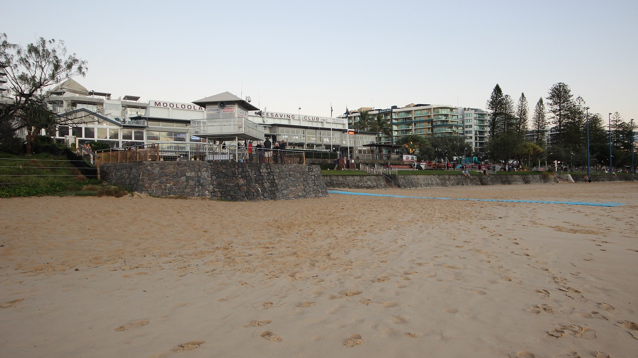 In the foreground, the sandy beach stretches out, showing footprints leading towards a stone wall that provides a boundary to the area. To the left, a grassy slope rises up to a raised platform, where a building labelled "MOLOOLABA" can be seen. This building features a deck with outdoor seating areas. On the right, additional structures are visible, including tall buildings surrounded by trees. In the foreground, the sandy beach stretches out, showing footprints leading towards a stone wall that provides a boundary to the area. To the left, a grassy slope rises up to a raised platform, where a building labelled "MOLOOLABA" can be seen. This building features a deck with outdoor seating areas. On the right, additional structures are visible, including tall buildings surrounded by trees.