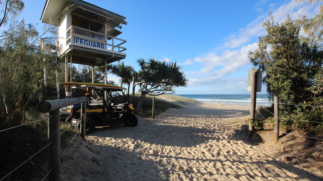To the left, there's a lifeguard tower elevated on stilts, marked by the word "LIFEGUARD" in bold letters. A metal ladder leads up to the platform. Nearby, an all-terrain vehicle is parked on the sandy path. On the right, there are clusters of beach vegetation, including palm trees and shrubs. The ground is covered in coarse sand, creating a distinct path leading towards the beach. There's no visible ceiling, as the area is open. To the left, there's a lifeguard tower elevated on stilts, marked by the word "LIFEGUARD" in bold letters. A metal ladder leads up to the platform. Nearby, an all-terrain vehicle is parked on the sandy path. On the right, there are clusters of beach vegetation, including palm trees and shrubs. The ground is covered in coarse sand, creating a distinct path leading towards the beach. There's no visible ceiling, as the area is open.