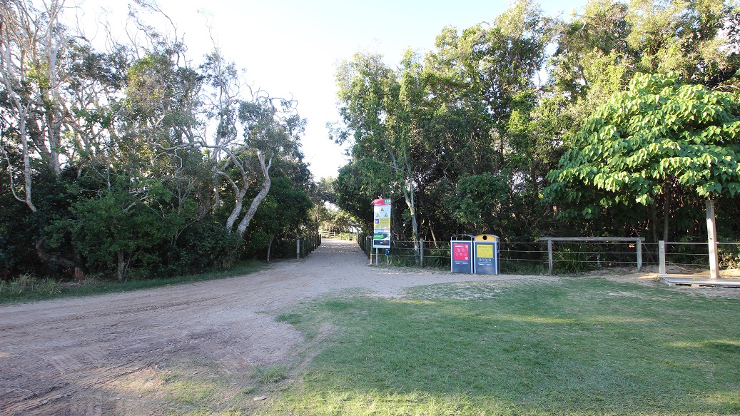 On the left, there's a pathway partially obscured by trees, indicating a wooded area. On the right, two recycling bins are positioned next to a signpost with colourful information boards. The ground is a dirt track with sparse grass, leading towards the trees in the background. The trees are dense, creating a natural border on either side of the path. There's no visible ceiling, as the area is outdoors. On the left, there's a pathway partially obscured by trees, indicating a wooded area. On the right, two recycling bins are positioned next to a signpost with colourful information boards. The ground is a dirt track with sparse grass, leading towards the trees in the background. The trees are dense, creating a natural border on either side of the path. There's no visible ceiling, as the area is outdoors.