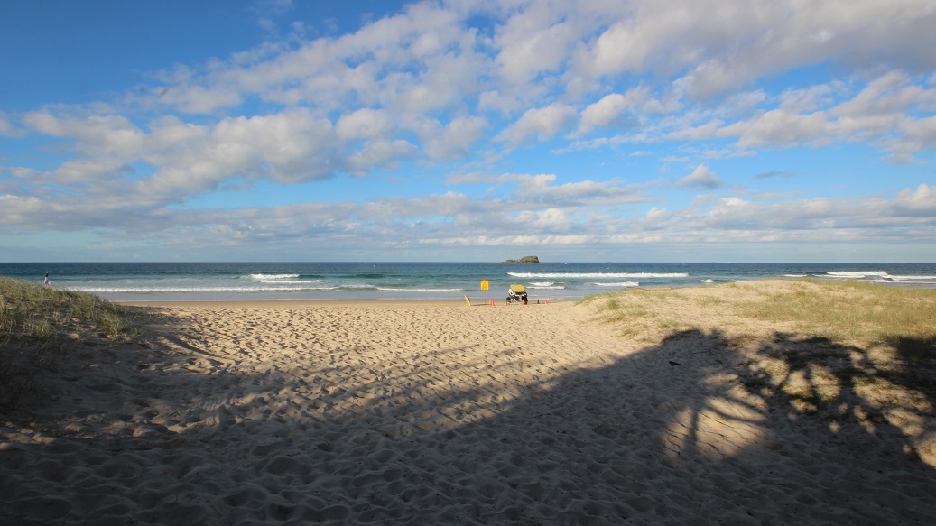 In the foreground, a sandy beach stretches out, with fine grains of sand visible. To the left, a small figure can be seen wading in the water. On the right, a lifeguard buoy is positioned near the shoreline. Beyond the beach, the ocean waves gently lap at the sand. In the distance, a rocky outcrop emerges from the water, adding interest to the horizon. There are patches of grass at the edge of the sand, providing a transition between the beach and the surrounding terrain. In the foreground, a sandy beach stretches out, with fine grains of sand visible. To the left, a small figure can be seen wading in the water. On the right, a lifeguard buoy is positioned near the shoreline. Beyond the beach, the ocean waves gently lap at the sand. In the distance, a rocky outcrop emerges from the water, adding interest to the horizon. There are patches of grass at the edge of the sand, providing a transition between the beach and the surrounding terrain.