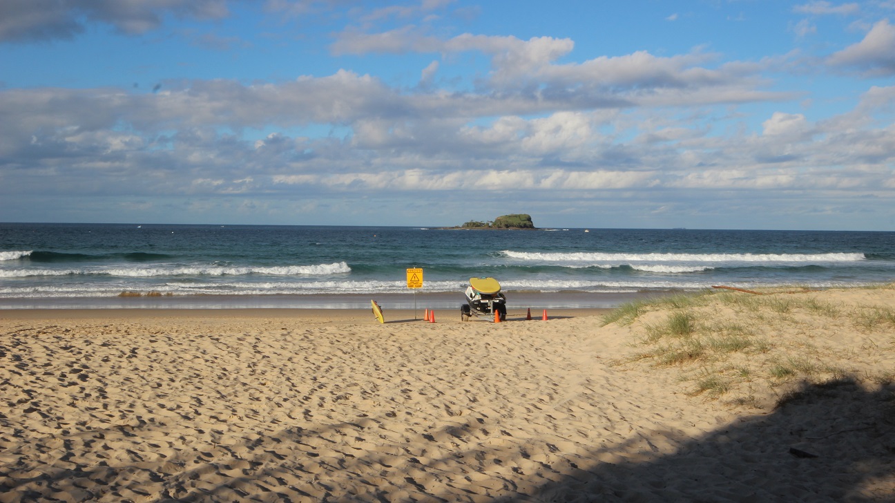 In the foreground, a sandy beach is visible, with fine sand and some grassy patches at the edges. To the right, a lifeguard vehicle is parked near a yellow warning sign, surrounded by orange traffic cones. In the middle, gentle waves roll onto the shore, creating a subtle separation between the sand and water. In the background, a rocky outcrop is situated offshore, providing a focal point against the water. The overall layout highlights the beach’s expanse, with no overhead structures in sight. In the foreground, a sandy beach is visible, with fine sand and some grassy patches at the edges. To the right, a lifeguard vehicle is parked near a yellow warning sign, surrounded by orange traffic cones. In the middle, gentle waves roll onto the shore, creating a subtle separation between the sand and water. In the background, a rocky outcrop is situated offshore, providing a focal point against the water. The overall layout highlights the beach’s expanse, with no overhead structures in sight.