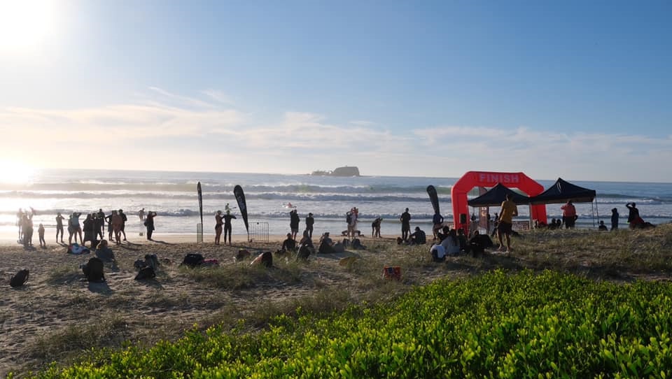 In the foreground, a group of people is scattered across the sandy beach, some sitting on the ground while others stand in small clusters. To the right, a bright red archway marked "FINISH" towers over a section with two black tents set up. There are several black flags nearby. The ground is sandy, with patches of grass and small shrubs visible. In the background, the ocean features rolling waves, and a rocky outcrop is visible on the horizon. In the foreground, a group of people is scattered across the sandy beach, some sitting on the ground while others stand in small clusters. To the right, a bright red archway marked "FINISH" towers over a section with two black tents set up. There are several black flags nearby. The ground is sandy, with patches of grass and small shrubs visible. In the background, the ocean features rolling waves, and a rocky outcrop is visible on the horizon.