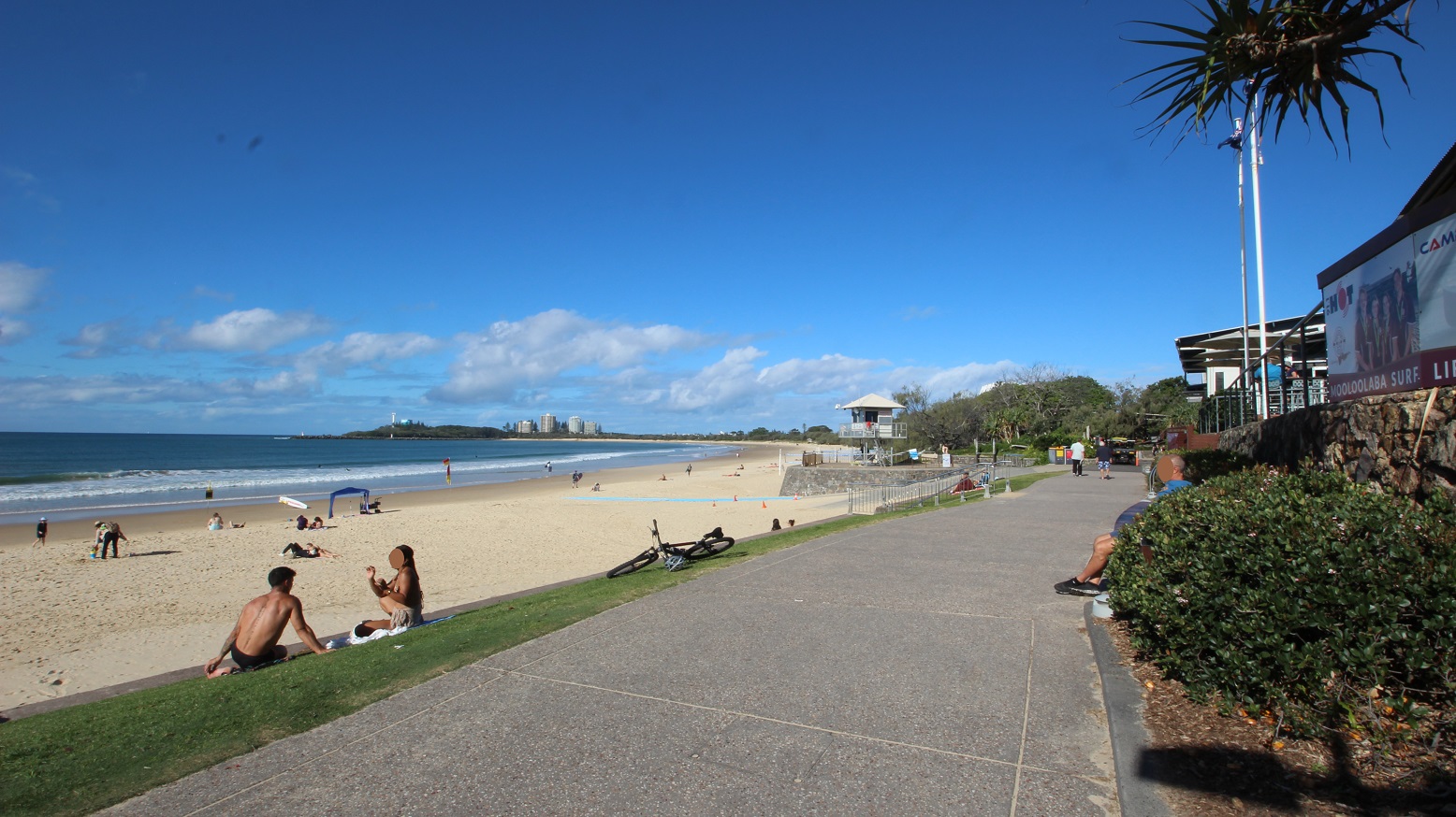 On the left, several people are relaxing on the sandy beach, with a couple sitting and others walking. To the right, a paved walkway runs alongside a grassy area, where individuals sit or stand. In the background, the beach stretches out towards the ocean, with a lifeguard tower visible, and tall buildings can be seen further back along the shoreline. The ground is a mix of smooth pavement on the walkway and soft sand on the beach. On the left, several people are relaxing on the sandy beach, with a couple sitting and others walking. To the right, a paved walkway runs alongside a grassy area, where individuals sit or stand. In the background, the beach stretches out towards the ocean, with a lifeguard tower visible, and tall buildings can be seen further back along the shoreline. The ground is a mix of smooth pavement on the walkway and soft sand on the beach.