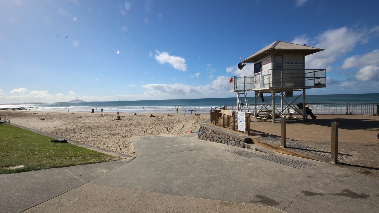 On the left, there is a grassy area adjacent to a paved walkway, with a few items laid out on the ground. In the centre, a lifeguard tower is elevated on stilts, providing a clear view of the beach and the ocean. To the right, there’s a spacious sandy beach where individuals are sunbathing and walking near the water’s edge. The floor is a mix of smooth pavement and soft sand, guiding visitors from the walkway to the beach. On the left, there is a grassy area adjacent to a paved walkway, with a few items laid out on the ground. In the centre, a lifeguard tower is elevated on stilts, providing a clear view of the beach and the ocean. To the right, there’s a spacious sandy beach where individuals are sunbathing and walking near the water’s edge. The floor is a mix of smooth pavement and soft sand, guiding visitors from the walkway to the beach.