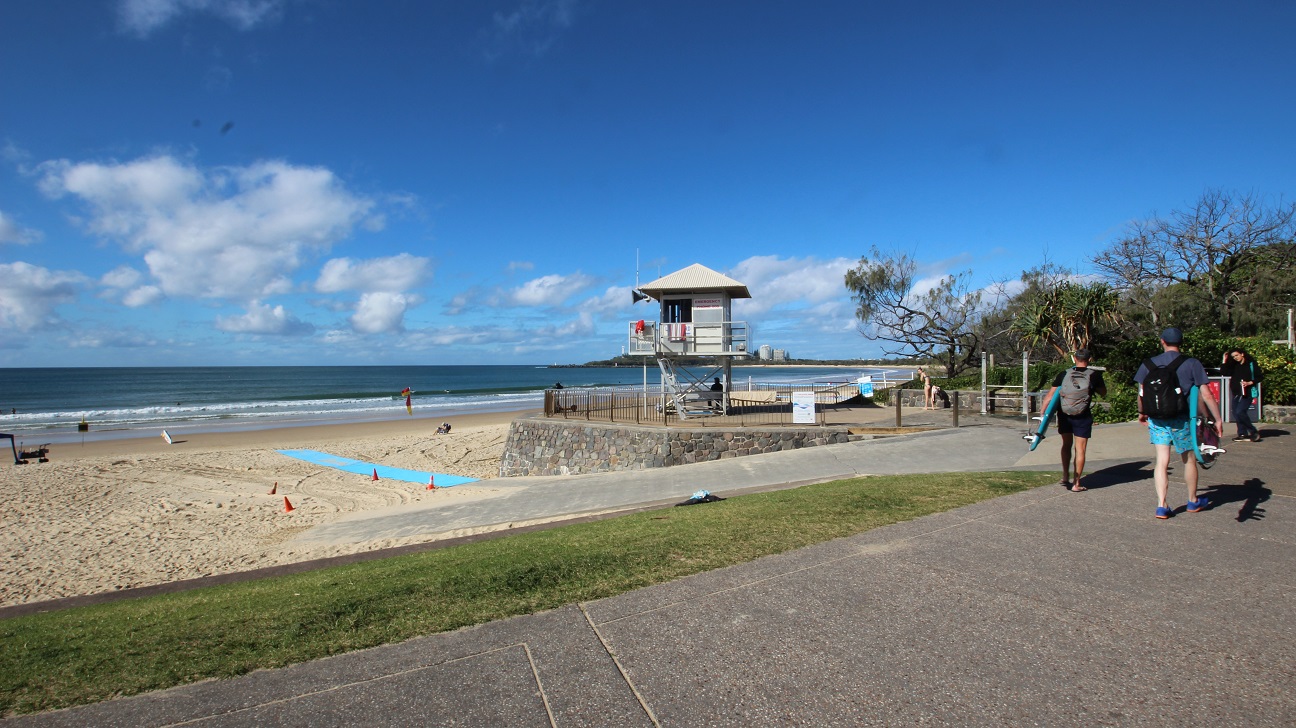 On the left, there is a sandy beach with a few people enjoying the sun, some sitting and others walking near the water's edge. Blue markers are placed on the sand, suggesting designated areas. In the centre, a lifeguard tower is positioned on elevated stilts, providing surveillance over the beach. To the right, two individuals are walking along the pathway, one carrying a surfboard. On the left, there is a sandy beach with a few people enjoying the sun, some sitting and others walking near the water's edge. Blue markers are placed on the sand, suggesting designated areas. In the centre, a lifeguard tower is positioned on elevated stilts, providing surveillance over the beach. To the right, two individuals are walking along the pathway, one carrying a surfboard.