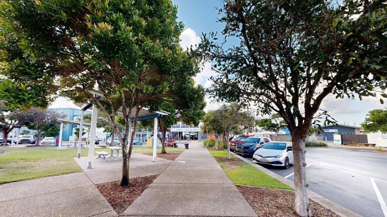 In the foreground, there are two symmetrical pathways lined with trees, creating a neat, shaded walkway. On the left, a seating area with white tables and benches is visible, alongside a few parked cars further down. On the right, additional parked vehicles are present, with a building fading into the background. The ground is paved with a grey concrete surface, and the area appears well-maintained. The scene has a clean, suburban feel, with greenery enhancing the walkways. In the foreground, there are two symmetrical pathways lined with trees, creating a neat, shaded walkway. On the left, a seating area with white tables and benches is visible, alongside a few parked cars further down. On the right, additional parked vehicles are present, with a building fading into the background. The ground is paved with a grey concrete surface, and the area appears well-maintained. The scene has a clean, suburban feel, with greenery enhancing the walkways.