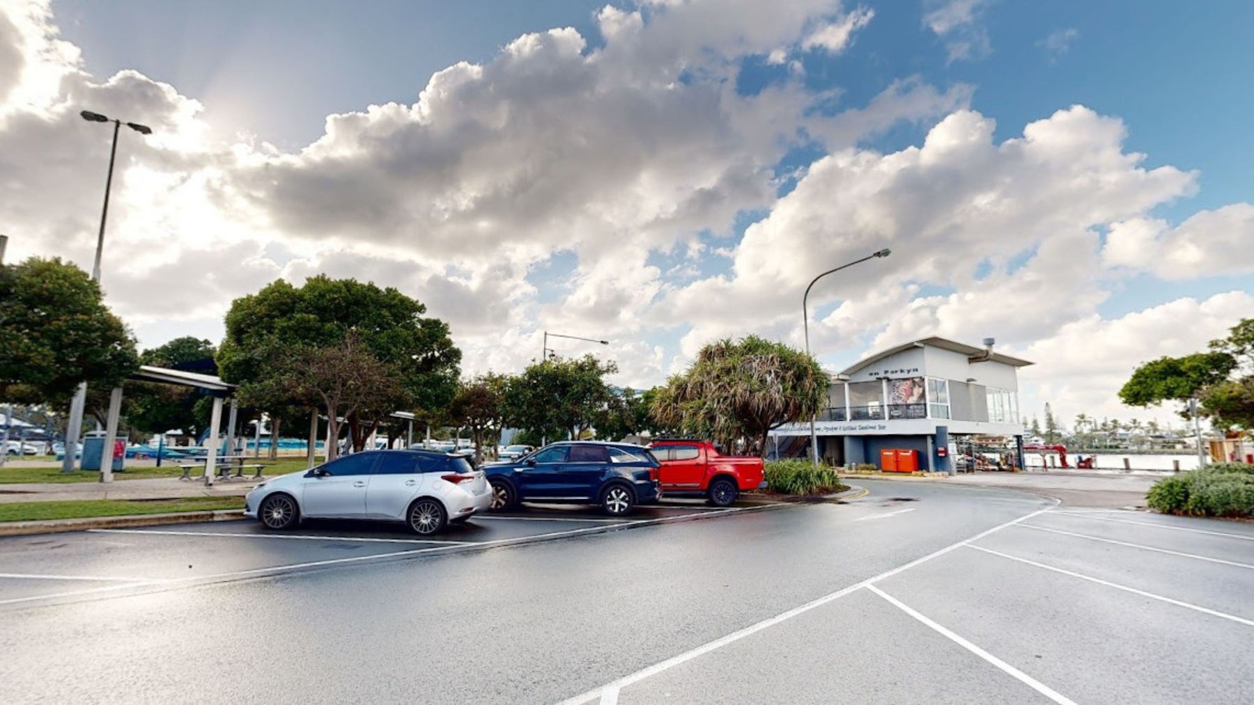 In the foreground, there are several parked cars, including a white sedan and a black hatchback, arranged along a smooth, wet asphalt surface. To the left, a seating area with covered benches is visible, surrounded by green trees. The background features a two-storey building with large windows, displaying a nautical theme and additional signage. A small group of trees is in front of the building, enhancing the area’s greenery. In the foreground, there are several parked cars, including a white sedan and a black hatchback, arranged along a smooth, wet asphalt surface. To the left, a seating area with covered benches is visible, surrounded by green trees. The background features a two-storey building with large windows, displaying a nautical theme and additional signage. A small group of trees is in front of the building, enhancing the area’s greenery.
