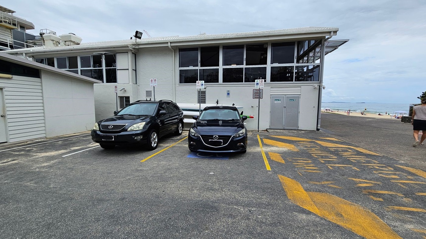 In the foreground, two cars are parked in marked spaces—a black SUV to the left and a black sedan to the right. The surface is paved with a grey concrete, with yellow markings indicating parking regulations. Directly behind the cars, a two-storey building features large windows, allowing visibility inside. To the left of the building, a smaller structure with a simpler facade is visible. In the foreground, two cars are parked in marked spaces—a black SUV to the left and a black sedan to the right. The surface is paved with a grey concrete, with yellow markings indicating parking regulations. Directly behind the cars, a two-storey building features large windows, allowing visibility inside. To the left of the building, a smaller structure with a simpler facade is visible.