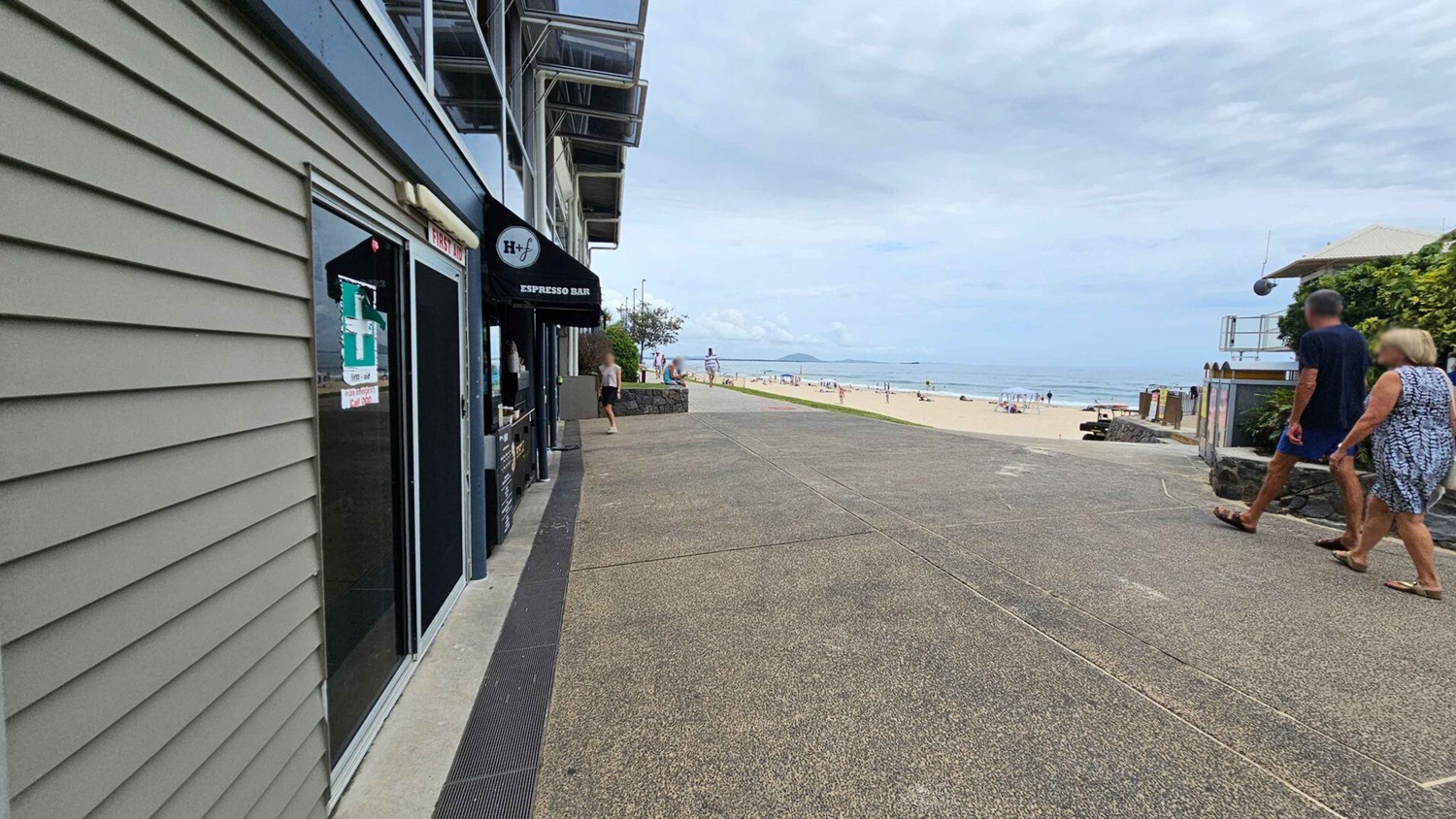 In the foreground, a wide concrete path leads towards the beach, with a textured surface and drainage channels on the sides. To the left, there is a building façade with large glass doors and a black awning labelled "Espresso Bar." On the right, a couple is walking along the path towards the beach. Beyond them, the beach is visible with people scattered across the sand. The pathway is well-maintained, providing a clear route to the shoreline. In the foreground, a wide concrete path leads towards the beach, with a textured surface and drainage channels on the sides. To the left, there is a building façade with large glass doors and a black awning labelled "Espresso Bar." On the right, a couple is walking along the path towards the beach. Beyond them, the beach is visible with people scattered across the sand. The pathway is well-maintained, providing a clear route to the shoreline.