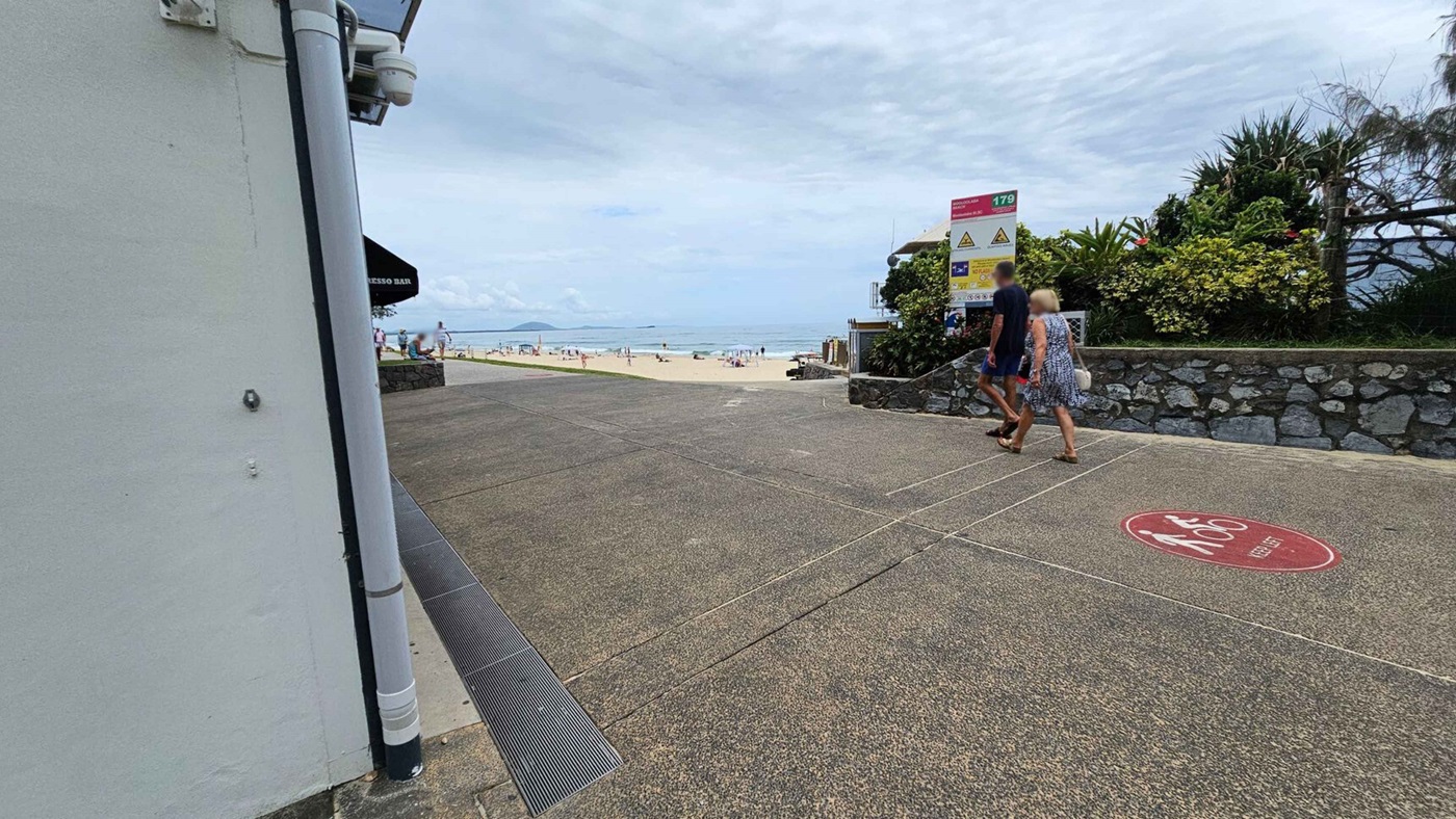 In the foreground, a broad concrete path leads toward the beach, marked with a circular red sign indicating a number. To the left, a white wall with a pipe is partially visible, and an awning from a nearby building provides shade. On the right, there’s a stone wall adorned with greenery, and beside it, a couple is walking towards the beach. In the background, the beach is visible with people enjoying the sand and surf, creating an atmosphere of leisure. In the foreground, a broad concrete path leads toward the beach, marked with a circular red sign indicating a number. To the left, a white wall with a pipe is partially visible, and an awning from a nearby building provides shade. On the right, there’s a stone wall adorned with greenery, and beside it, a couple is walking towards the beach. In the background, the beach is visible with people enjoying the sand and surf, creating an atmosphere of leisure.